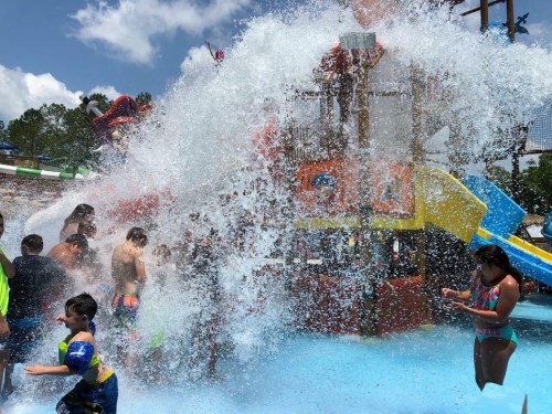A water park scene with kids splashing in a pool, a large water slide, and a big splash from a water feature.
