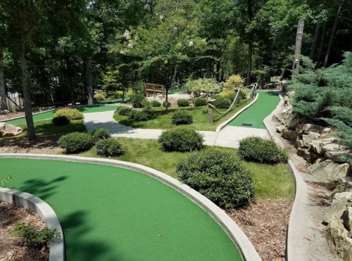 A scenic mini-golf course with green turf, winding paths, and lush landscaping surrounded by trees.