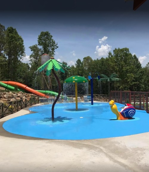 A colorful water play area featuring a snail slide, palm trees, and a large water splash feature under a blue sky.