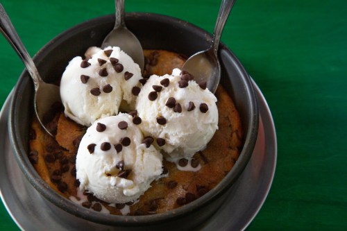 A warm chocolate chip cookie topped with three scoops of vanilla ice cream and chocolate chips, served with spoons.