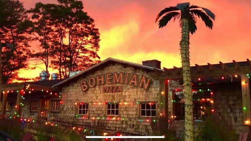 A cozy pizza restaurant with a wooden exterior, adorned with colorful lights, against a vibrant sunset sky.