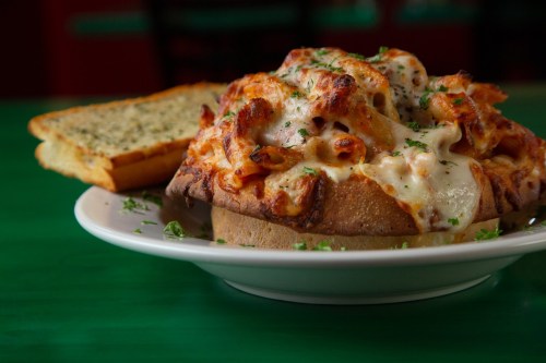 A cheesy pasta dish served in a bread bowl, garnished with parsley, alongside a slice of garlic bread.