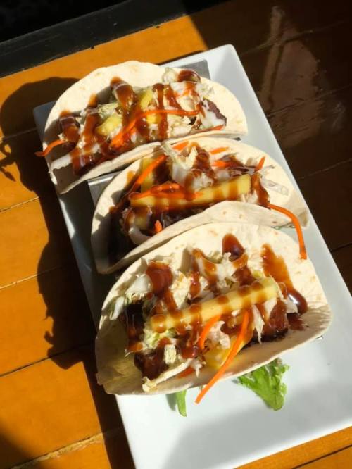 Three tacos on a plate, filled with shredded meat, cheese, and topped with sauce and fresh vegetables.