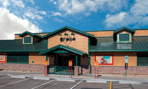 Exterior of a restaurant with a green roof and wooden accents, featuring a sign and parking area under a blue sky.