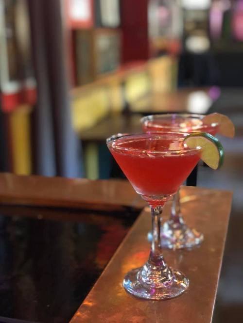 Two vibrant red cocktails in elegant glasses, garnished with lime, sit on a bar counter with a blurred background.