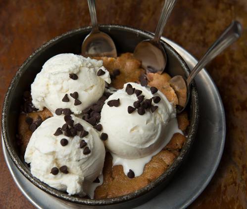 A warm cookie dessert topped with three scoops of vanilla ice cream and chocolate chips, served in a metal bowl.