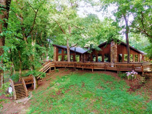 A modern wooden house surrounded by lush greenery and trees, featuring a deck and stairs leading to the ground.