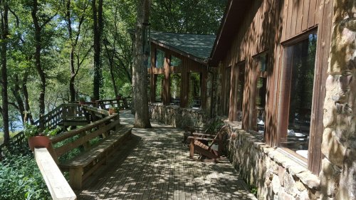 A wooden deck surrounds a rustic cabin, nestled among trees by a river, with large windows and stone accents.