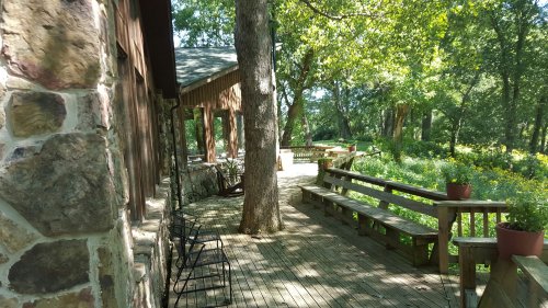 A wooden deck with benches, surrounded by trees and flowers, next to a stone building in a serene outdoor setting.