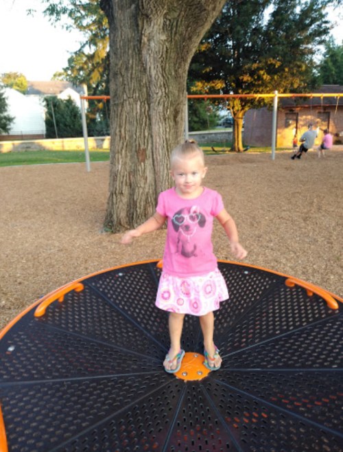 A young girl in a pink shirt and skirt stands on a spinning playground structure, smiling in a park setting.