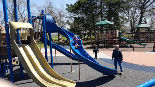 A child slides down a blue slide at a playground, while an adult and another child play nearby on a sunny day.
