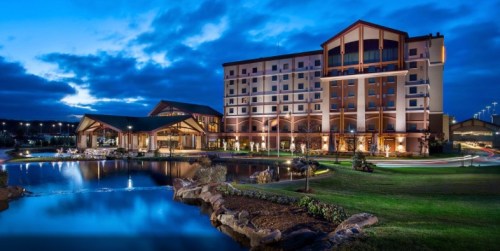 A modern hotel at dusk, featuring a pond, landscaped grounds, and illuminated architecture against a cloudy sky.