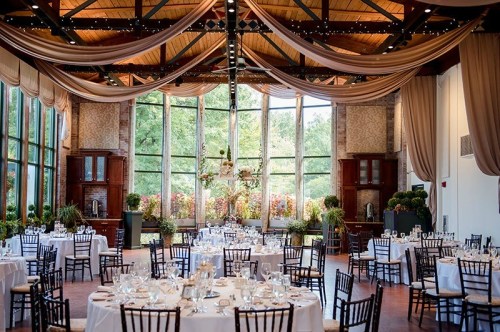 Elegant event space with round tables, white linens, and large windows showcasing greenery outside.
