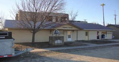 A single-story community building with a bell and a tree in front, set in a rural area.
