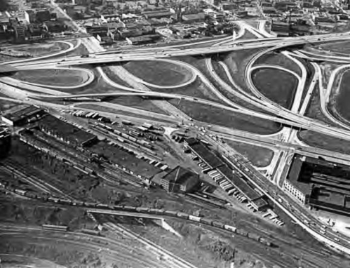 Aerial view of a complex highway interchange with roads, rail tracks, and buildings in a cityscape.