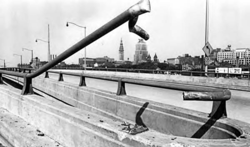 A broken metal railing on a highway overpass, with a city skyline in the background, captured in black and white.