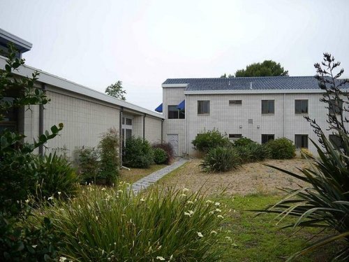 A modern building surrounded by greenery and a gravel pathway, under a cloudy sky.