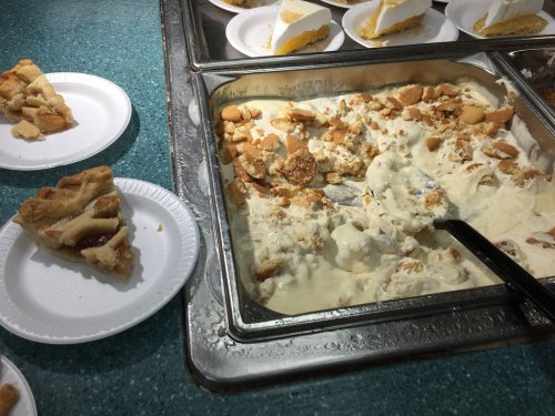 A tray of banana pudding topped with crushed cookies, next to a slice of pie on a paper plate.