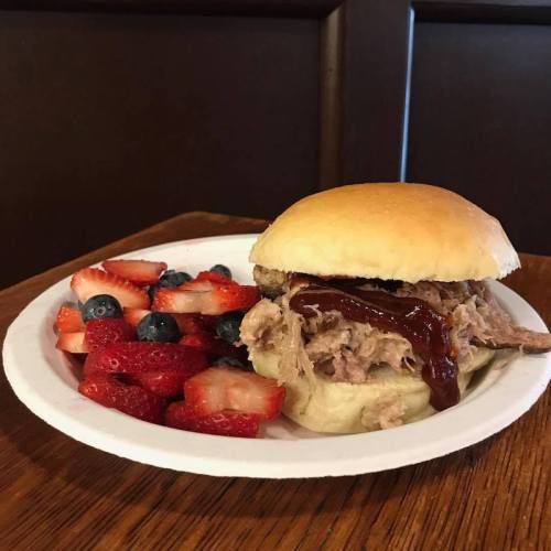 A pulled pork sandwich with barbecue sauce on a bun, served with a side of strawberries and blueberries.