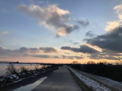 A serene pathway beside a river, with snow on the ground and a colorful sunset sky filled with clouds.