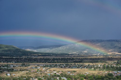 A vibrant rainbow arcs over a mountainous landscape, with a valley and small town visible below.