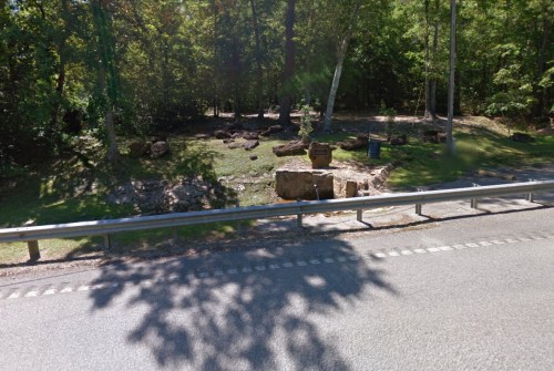 A wooded area beside a road featuring a rock formation and a sign, with trees and greenery in the background.