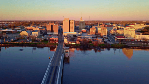Aerial view of a city skyline at sunset, reflecting on a river with a bridge in the foreground.