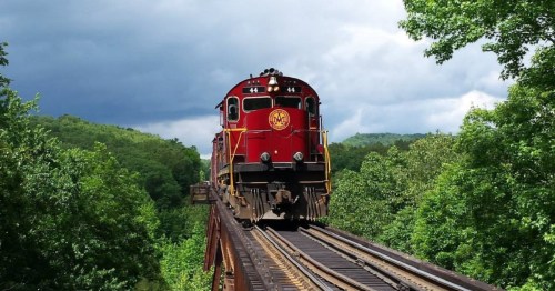 A red train crosses a bridge surrounded by lush green trees under a cloudy sky.