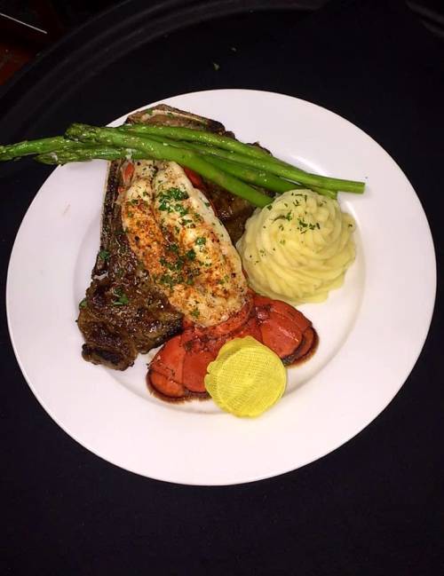 A gourmet plate featuring grilled lobster, steak, mashed potatoes, asparagus, and a slice of lemon on a black background.