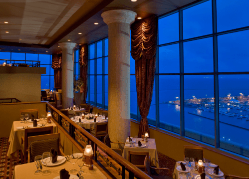 Elegant restaurant interior with tables set for dining, large windows showcasing a nighttime waterfront view.