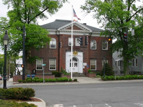 Historic brick building with a flag, surrounded by trees, located at a street intersection with traffic lights.