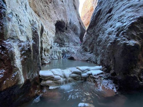 A narrow canyon with water pooling around sandbags, surrounded by rocky walls and sunlight filtering through above.