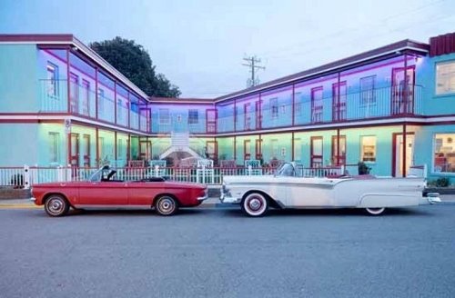 Two classic convertible cars parked in front of a colorful motel with vibrant lighting at dusk.