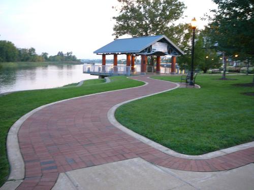 A winding brick path leads to a gazebo by a calm river, surrounded by green grass and trees at dusk.