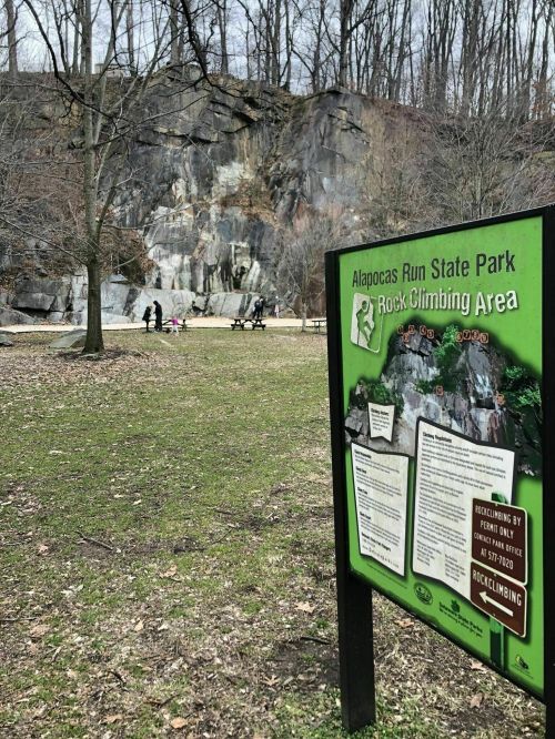 Sign for the rock climbing area at Alapocas Run State Park, with a rocky cliff and people in the background.