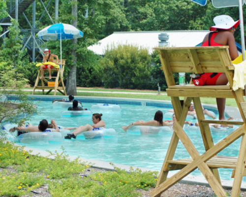 A lifeguard watches over a pool where people float on tubes, surrounded by trees and umbrellas.