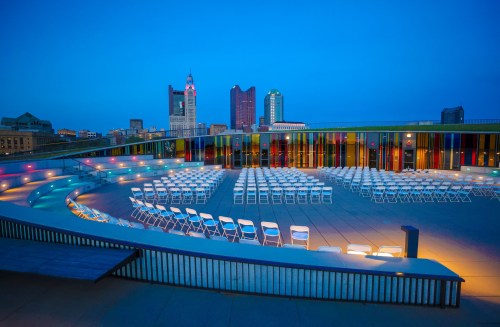 A rooftop venue set up with rows of white chairs, city skyline in the background, illuminated at dusk.