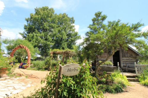 A garden scene featuring a "Herbs" sign, a rustic cabin, and lush greenery under a blue sky.