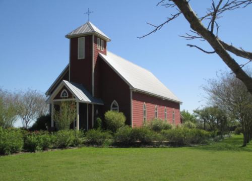 A red church with a steeple, surrounded by green grass and trees under a clear blue sky.