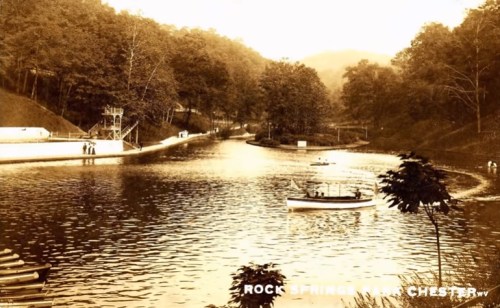 A vintage photo of a serene river scene with boats, surrounded by trees and hills.