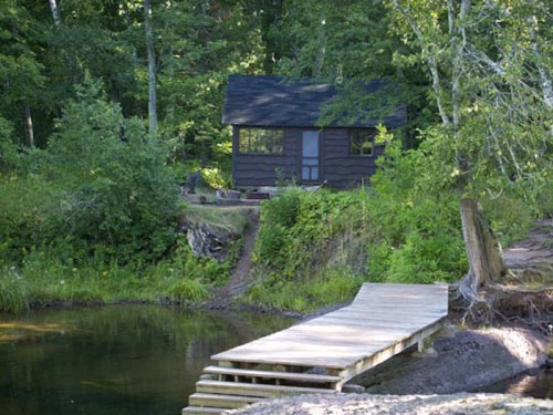A rustic cabin surrounded by lush greenery, with a wooden dock leading to a calm pond.