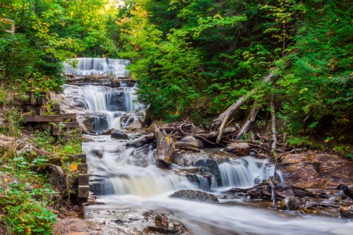 A serene waterfall cascades over rocks, surrounded by lush green trees and vibrant autumn foliage.