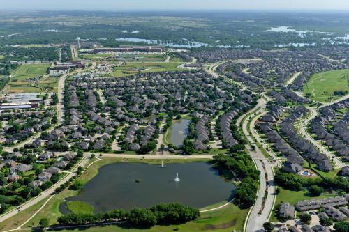 Aerial view of a suburban neighborhood with houses, a pond, and green spaces under a clear blue sky.