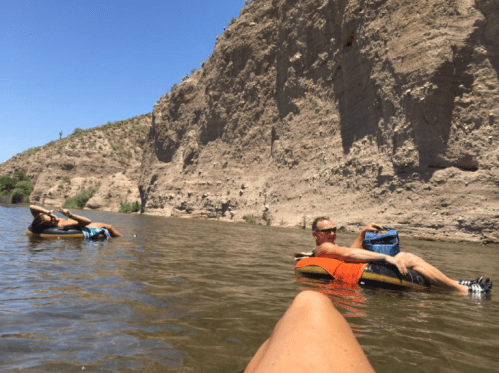 Two people float on inner tubes in a river, surrounded by rocky cliffs and clear blue skies.
