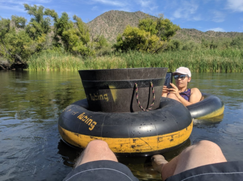 A person relaxes on a tube in a river, using a phone, with mountains and greenery in the background.