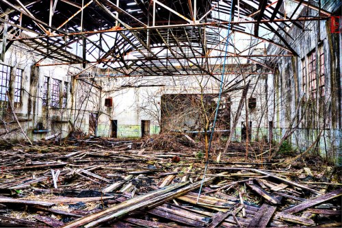 An abandoned building with a collapsed roof, overgrown with plants and scattered wooden debris on the floor.