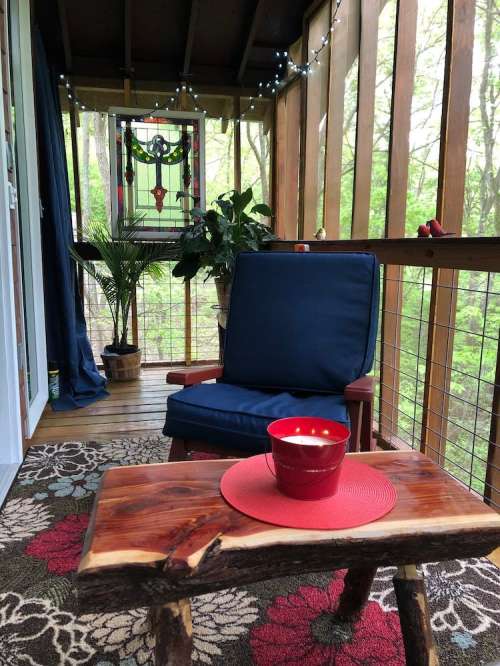 Cozy porch with a blue chair, wooden table, red candle, plants, and stained glass window, surrounded by greenery.