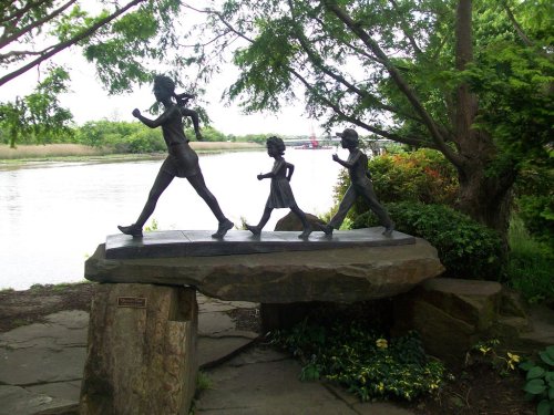 A bronze sculpture of a woman and two children running along a riverbank, surrounded by greenery.