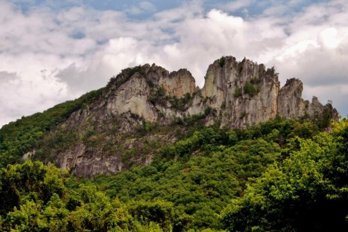 A rugged mountain peak rises above lush green trees under a partly cloudy sky.