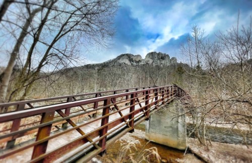 A wooden bridge spans a stream, with rocky hills and a cloudy sky in the background, surrounded by bare trees.